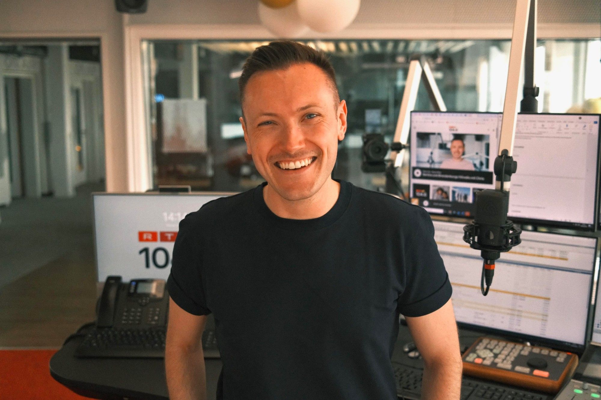 Chris Hartmann lachend im Sendestudio von 104.6 RTL, hinter ihm Mikrofon und Bildschirme